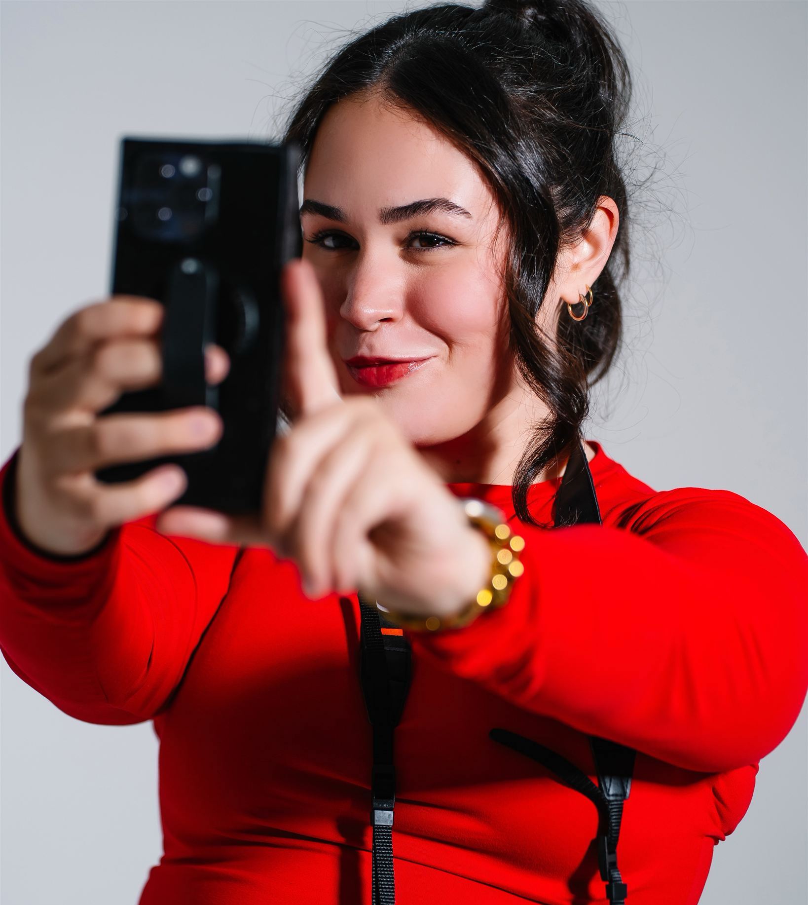 Sophia Pearle in a red top, smiling as she holds a phone up to film and points toward the camera, against a grey studio backdrop.