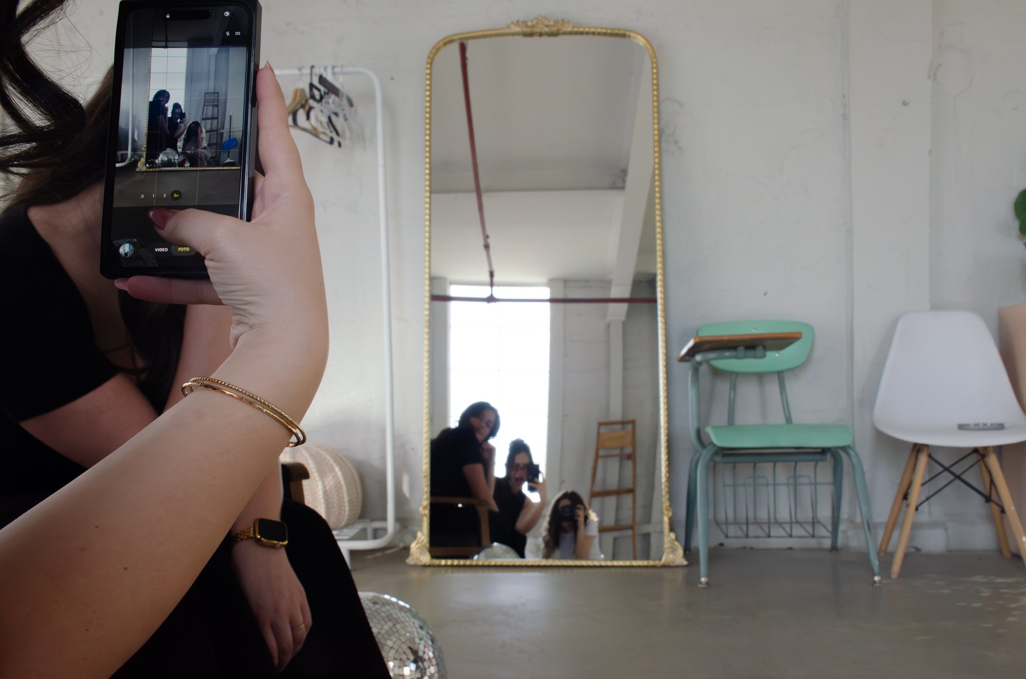 Sophia and two clients reflected in a tall gilded floor mirror, laughing as one of them takes a photo.