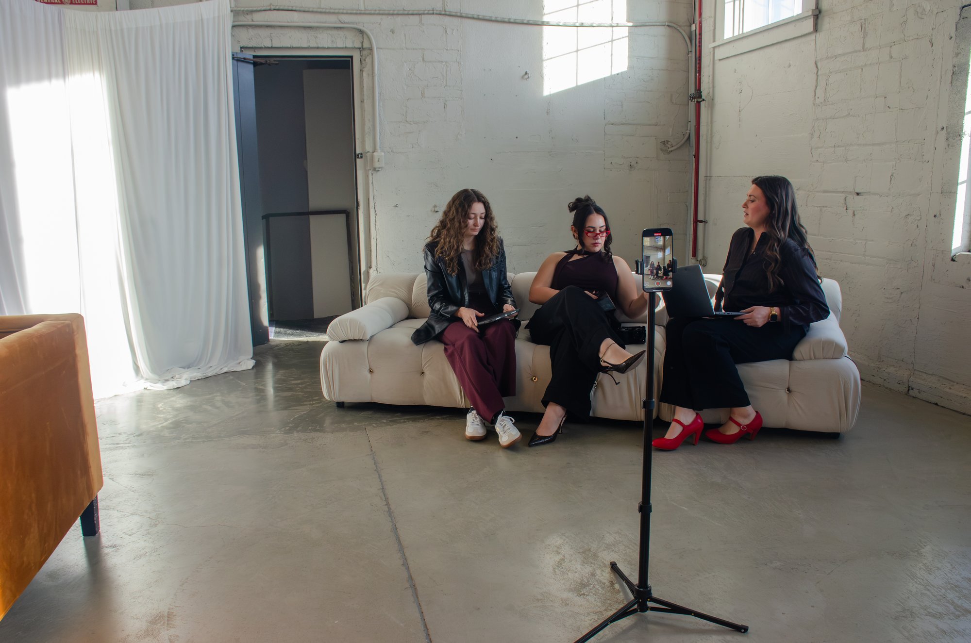 A wide view of a warehouse studio: three people on a cream sofa filming a seated interview with a phone on a tripod.