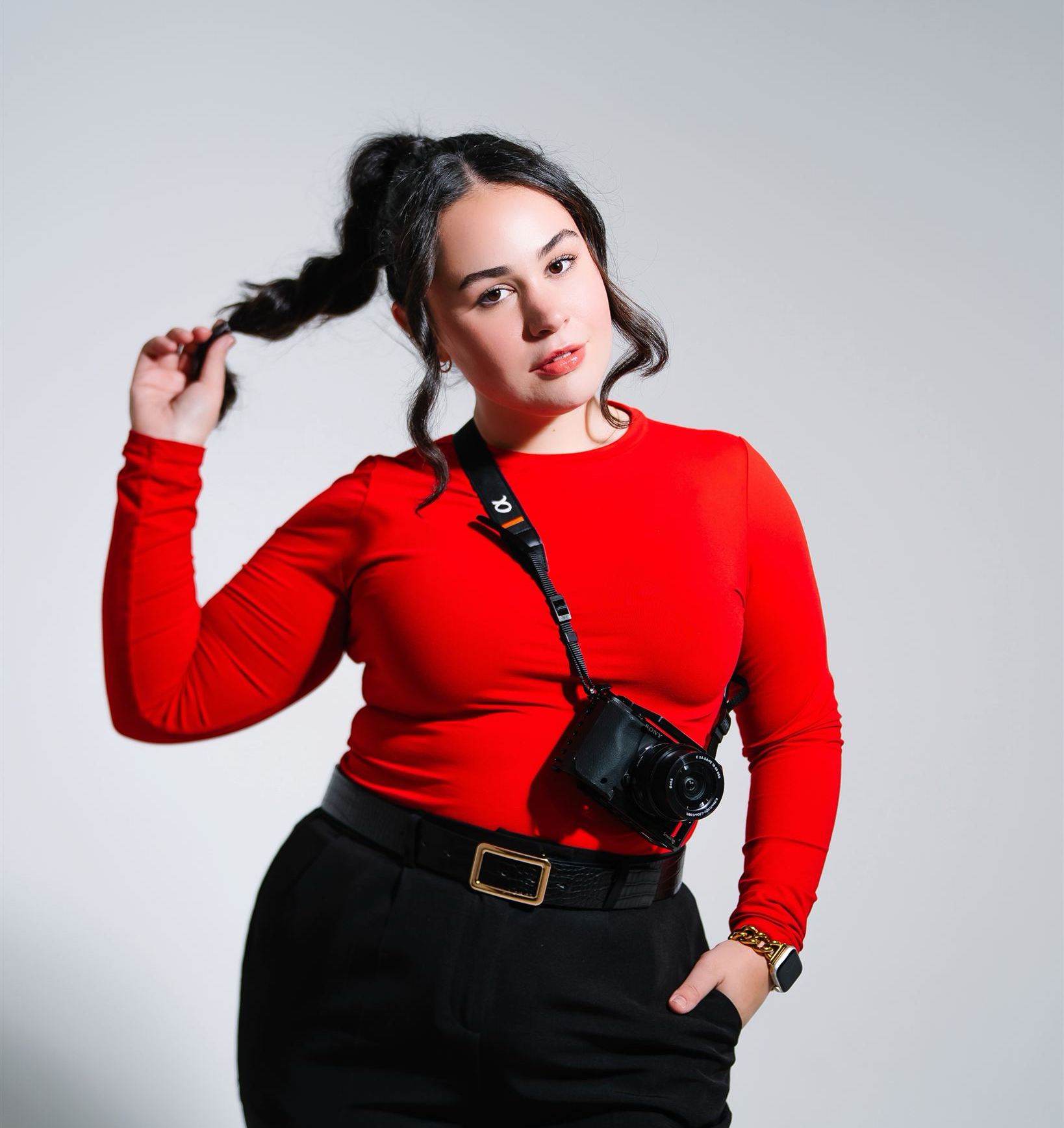Sophia Pearle in a red long-sleeve top with a camera around her neck, one hand tugging her ponytail, against a grey studio backdrop.