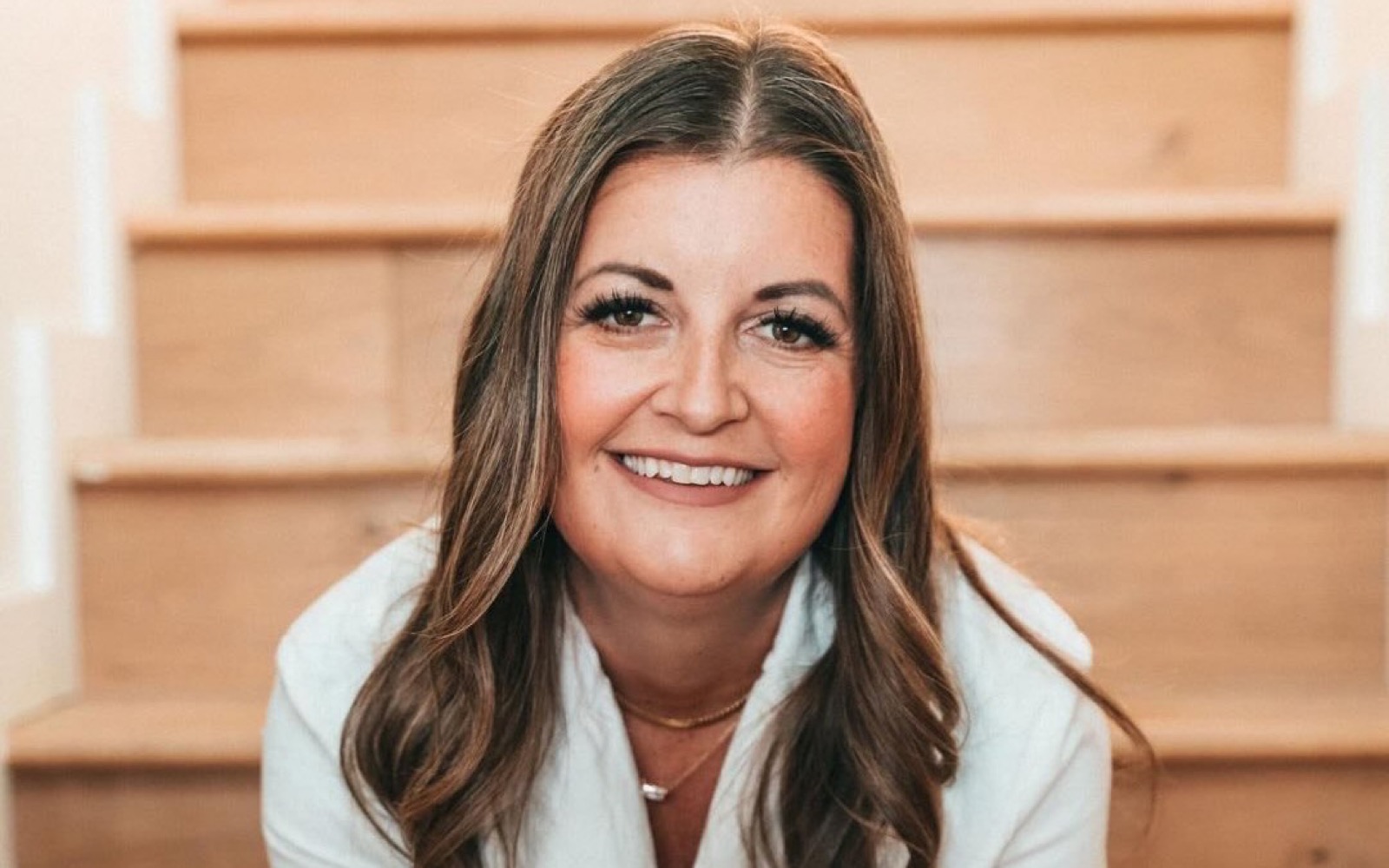 Jess Lay, a real estate agent in a cream blazer, smiling while seated on wooden stairs.