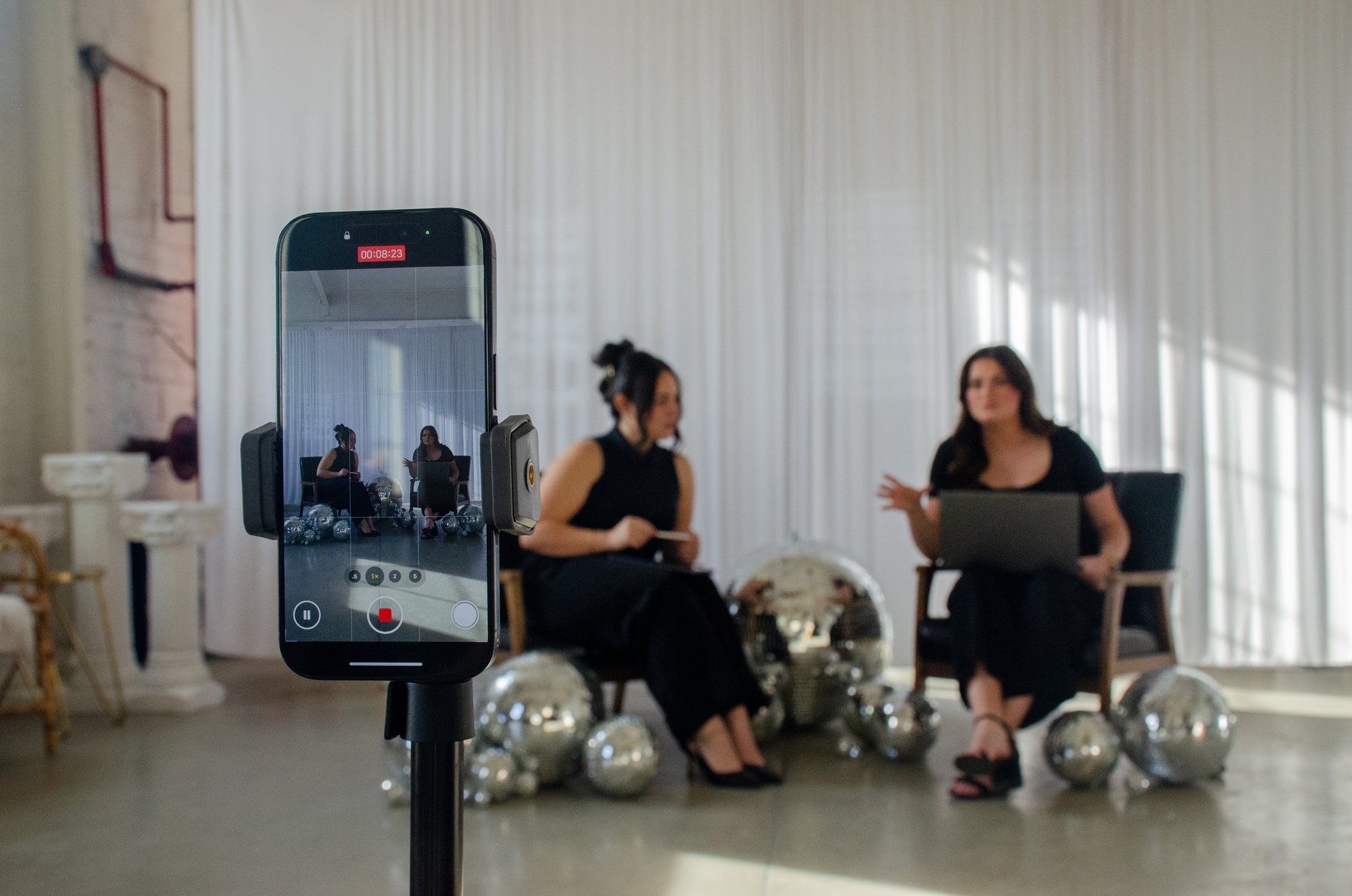 A phone on a tripod recording two people mid-conversation on a bright studio set dressed with disco balls.