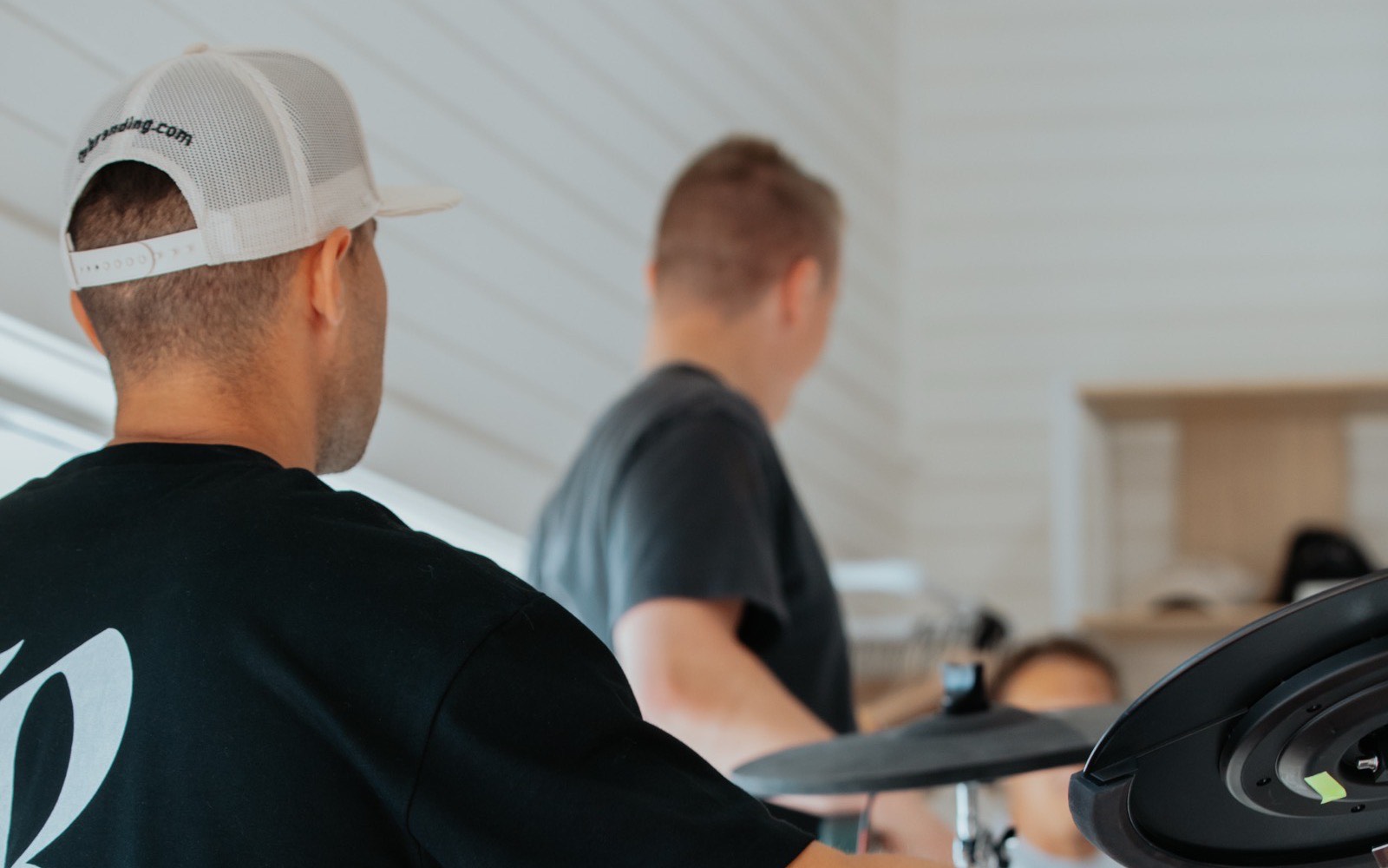 A worship band mid-practice, a drummer in a black Rise tee at an electronic kit with a guitarist and keyboardist behind.