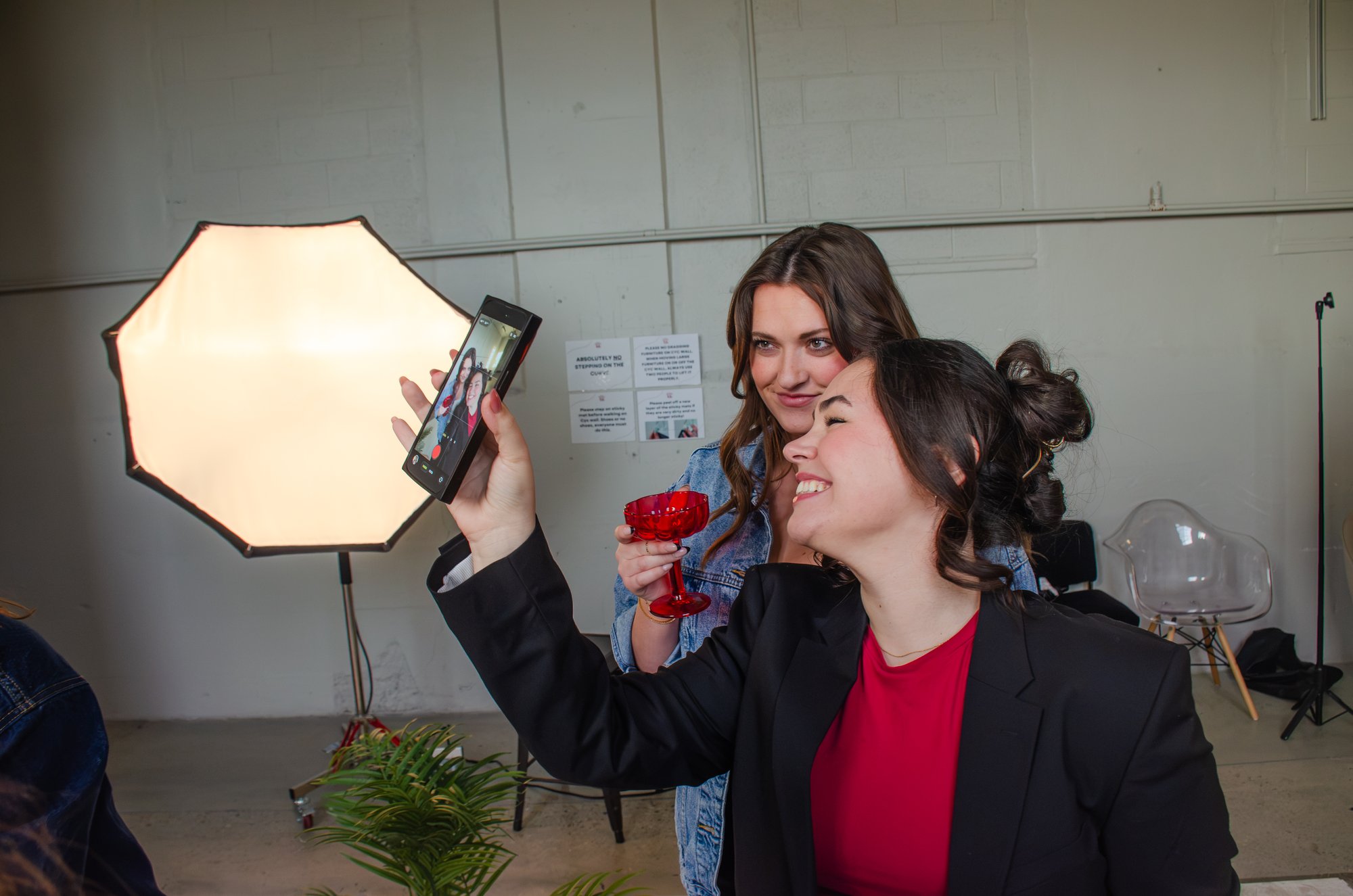 Two people laughing while filming a selfie video together on a studio set, a softbox light glowing behind them.